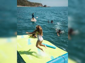 A group enjoys a fun day on the water, with a child jumping off a floating mat while others paddleboard and swim nearby — perfect for summer adventures.