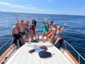 A lively group of friends celebrates on the bow of a boat under the sun, showcasing the joy of summer outings on the water.