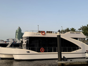 A sleek yacht named 'Dudu Yacht' docked at the marina, showcasing its modern design against a backdrop of palm trees and urban architecture.