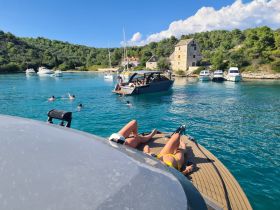 A lively scene captures guests enjoying a sunny day by the water, swimming and sunbathing on a pontoon boat, surrounded by beautiful scenery and other boats.