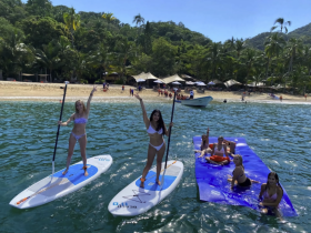 A lively group enjoys paddleboarding and relaxing on a floating mat at a beautiful beach, showcasing the perfect day of fun in the sun.