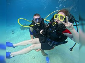 A couple enjoys an underwater diving adventure, showcasing their excitement with a peace sign while exploring vibrant marine life.