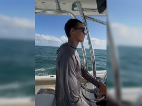 A young man navigates the helm of a boat, showcasing the interior console and the beautiful ocean backdrop — perfect for those looking to experience boating adventures.