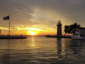 A stunning sunset view at the harbor, featuring a lighthouse and a boat, with people enjoying the picturesque scenery — perfect for romantic evenings or scenic cruises.