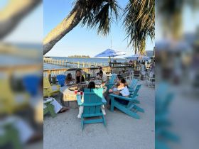 A lively group enjoys outdoor dining around a fire pit, surrounded by colorful chairs and scenic waterfront views — perfect for gatherings and celebrations.