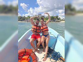 A joyful couple poses on a small boat near Tulum, showcasing their adventure with life jackets and a stunning backdrop of ancient ruins — perfect for memorable experiences on the water.
