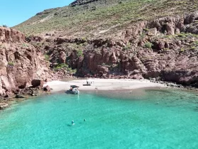A stunning aerial view of a secluded beach with crystal-clear waters, where guests enjoy swimming and relaxation near a boat — an ideal destination for a day of adventure and exploration.
