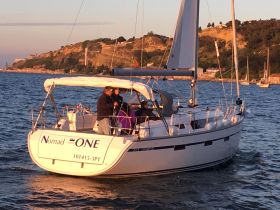 A couple enjoys a sailing experience aboard the 'Nomad ONE' as the sun sets, creating a picturesque backdrop in the harbor.