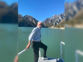 A man poses on the deck of a boat with stunning mountain scenery and a serene lake in the background, capturing the essence of a perfect day on the water.