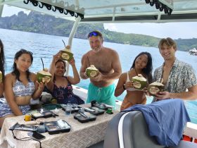 A cheerful group enjoys refreshing coconuts and a meal aboard a boat, surrounded by stunning coastal scenery — perfect for a fun day on the water.