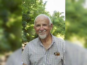 A smiling older man poses for a photo in a lush green setting, capturing a moment of joy amidst nature.