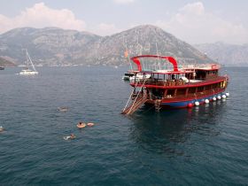 A vibrant scene of guests enjoying a swim near a traditional boat, surrounded by stunning mountainous coastline — perfect for a day of adventure on the water.