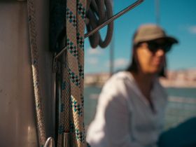 A partially blurred image featuring a person on a boat, with ropes in focus. The unclear angle makes it difficult to discern details.