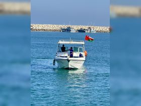 A small boat navigates through clear waters, showcasing a vibrant flag and two individuals onboard, perfect for exploring coastal destinations.