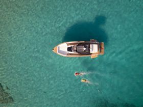 An aerial view captures two swimmers enjoying the clear turquoise waters near a stylish boat, showcasing the perfect day for water activities.