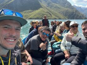 A lively group enjoys a boat trip, smiling and giving thumbs up against a stunning mountainous backdrop — perfect for adventure seekers and group outings.