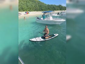 A man enjoys paddleboarding in crystal-clear waters, surrounded by friends and boats at a beautiful beach — perfect for water sports and relaxation.
