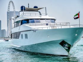 A stunning yacht cruising through clear waters, showcasing its sleek design against a backdrop of modern architecture and a prominent Ferris wheel.