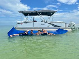 A fun group enjoys a day on the water, lounging on a floating mat next to a pontoon boat — perfect for family outings and water activities.