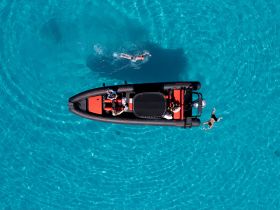 An aerial view captures a group enjoying a day on the water, with a boat anchored nearby and swimmers in the vibrant turquoise sea — perfect for summer adventures.