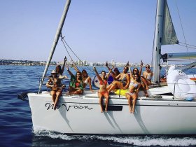 A lively group of friends enjoys a sailing adventure on the yacht 'My Vision,' celebrating with drinks and laughter against a stunning coastal backdrop.