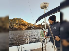A man enjoys the scenic view from a sailboat, with a picturesque coastline and lighthouse in the background — perfect for sunset cruises and coastal explorations.