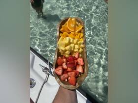 A vibrant platter of fresh fruit, including strawberries, mango, pineapple, and oranges, perfect for enjoying on a boat trip. A person is partially visible in the background, adding to the lively atmosphere.