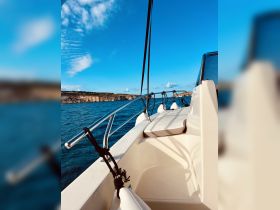 A serene view of the side of a boat cruising through clear waters, showcasing the vessel's sleek design against a backdrop of blue skies and distant cliffs.
