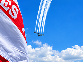 Aerial display of jets performing maneuvers against a bright blue sky, with a flag in the foreground. The focus is on the aircraft, creating a dynamic scene.