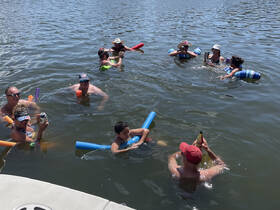 A lively group enjoys a fun day in the water, complete with pool noodles and drinks, making for a perfect summer party atmosphere.