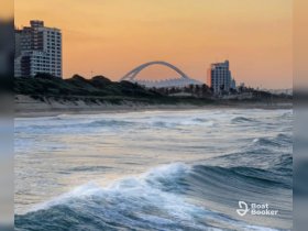 A scenic view of waves crashing against the shore at sunset, with buildings in the background, creating a tranquil coastal atmosphere.