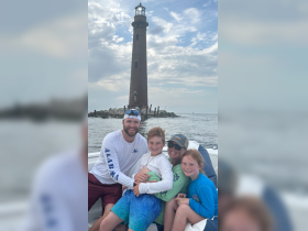 A joyful family poses on their boat with a lighthouse in the background, capturing a memorable moment on the water.
