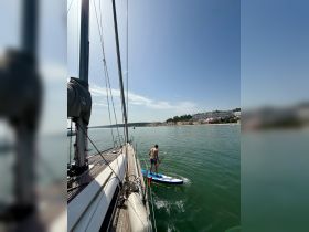 A man paddleboards alongside a sailboat, enjoying a sunny day on the water — a perfect summer activity for adventure seekers.