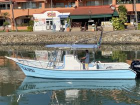 A fishing boat named 'La Otra' navigates the calm waters, with a captain at the helm, showcasing the vibrant marina atmosphere.
