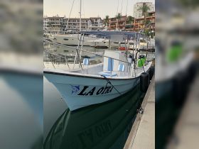 A well-maintained fishing boat named 'LA OTRA' docked in a marina, reflecting the serene surroundings in the calm waters.