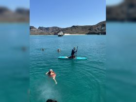 A lively scene featuring guests enjoying swimming and paddleboarding in clear turquoise waters, with a boat anchored in the background — perfect for a fun day on the water.