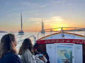 Two guests enjoy a scenic sunset view from the deck of a boat, with other sailboats in the background, creating a perfect atmosphere for a memorable sailing experience.
