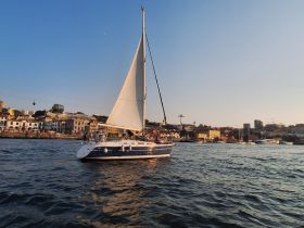 A beautiful sailboat glides through the water, showcasing its elegant sails against a vibrant backdrop of coastal buildings.
