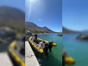 A vibrant yellow speedboat is docked at the shore, with a person preparing equipment against a stunning backdrop of mountains and clear waters.