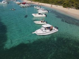A vibrant scene of boats anchored near a sandy beach, with groups enjoying the water and sun — a perfect spot for a sandbar stop and fun on the water.