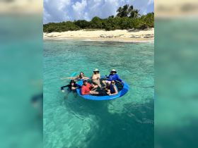 A lively group enjoys a relaxing day in the water on a floating mat, surrounded by stunning beach scenery — perfect for fun and socializing under the sun.