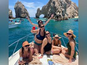 A lively group of friends enjoys a fun day on the water in Cabo San Lucas, celebrating with drinks and laughter aboard their boat, with stunning rock formations in the background.