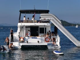 A lively group enjoys a day on the water aboard a catamaran, featuring a fun slide and paddleboarding activities — perfect for summer adventures.