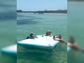 A group of friends enjoys a sunny day on a floating mat in clear turquoise waters, perfect for relaxation and fun in the sun.