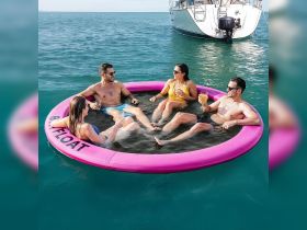 A lively group enjoys drinks while relaxing on a floating mat in the water, with a boat anchored nearby — perfect for summer fun and social gatherings on the water.
