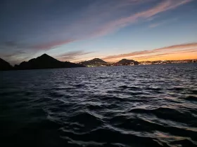 A stunning sunset view over the coastline, with silhouettes of mountains and city lights reflecting on the water — perfect for capturing the beauty of evening boat trips.