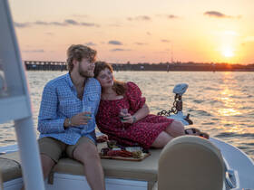 A couple enjoys a romantic sunset cruise on a boat, sipping cocktails and sharing a platter of snacks — the perfect setting for special moments on the water.
