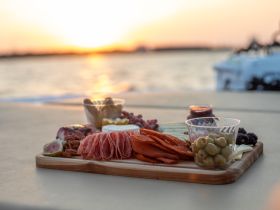 A beautifully arranged charcuterie board featuring a variety of meats, cheeses, and fruits, set against a stunning sunset backdrop — perfect for enhancing any boating experience.