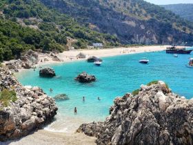 A stunning view of a beach with crystal-clear waters, rocky formations, and boats anchored nearby, showcasing a perfect summer day for swimming and relaxation.