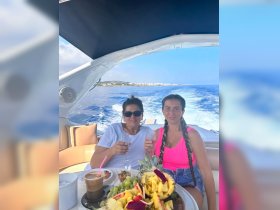 Two women enjoy cocktails and a vibrant fruit platter aboard a yacht, celebrating their time on the water with stunning views in the background.
